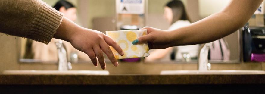 Yellow and white menstrual pad being passed between two people in a bathroom